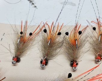 May include: Four brown and orange shrimp fly fishing lures with black beads for eyes. The lures have a brown and white feather body and orange legs.