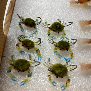 May include: Six green and yellow crab-shaped fishing lures with blue accents and black eyes. The lures are arranged in a row on a white foam surface.