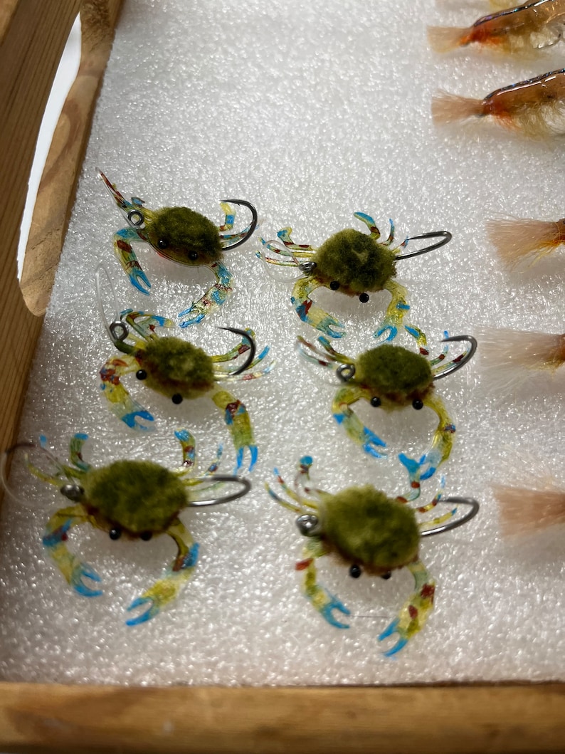 May include: Six green and yellow crab-shaped fishing lures with blue accents and black eyes. The lures are arranged in a row on a white foam surface.