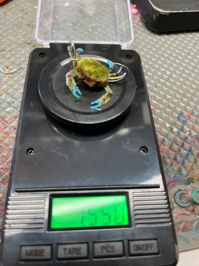 May include: A small, green and blue crab lure, weighing 1.55 grams, on a digital scale.