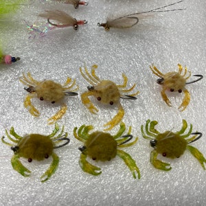 May include: Six realistic-looking crab fly fishing lures. Three are brown with yellow accents and three are green with yellow accents. The lures are on a white surface.