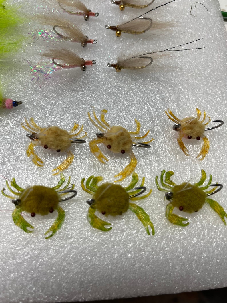 May include: Six realistic-looking crab fly fishing lures. Three are brown with yellow accents and three are green with yellow accents. The lures are on a white surface.