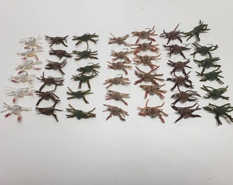 May include: A collection of 48 realistic-looking crab fishing lures in various colours, including white, brown, and green. The lures are arranged in rows on a white surface.