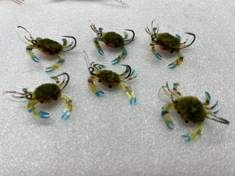 May include: Six green and brown crab-shaped fishing lures with clear plastic bodies, blue and white accents, and silver hooks.