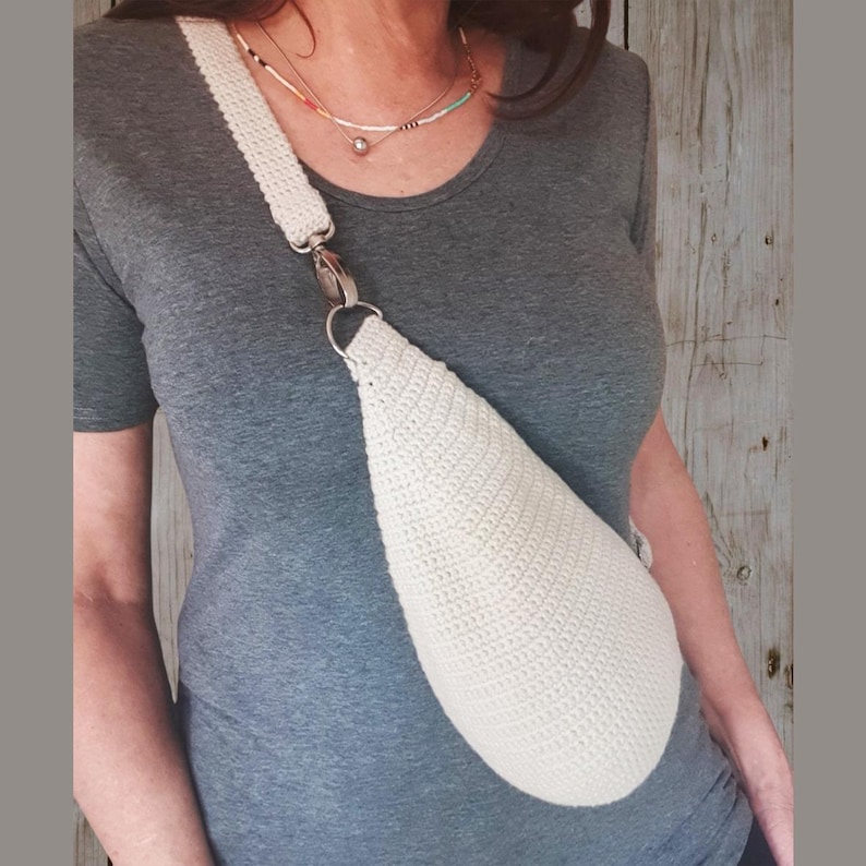 Puede incluir: Un bolso bandolera de crochet blanco con cierre plateado. El bolso se lleva sobre el hombro de una persona que lleva una camisa gris.