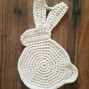 May include: A cream-colored, crocheted bunny-shaped trivet or potholder. The bunny has long ears and a circular body, with a textured, handmade appearance. The trivet is designed to protect surfaces from heat.