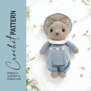Może przedstawiać: Szydełkowa lalka kota w niebieskim kombinezonie i pasującym kapeluszu. Lalka ma beżową głowę i łapy, różowy nos i czarne oczy. Tekst "Crochet Pattern" i "Pattern is available in English only" jest widoczny.