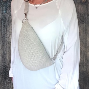 Puede incluir: Un bolso bandolera de crochet gris claro con una correa de cadena plateada. El bolso se lleva sobre el hombro de una persona que lleva una camisa blanca.