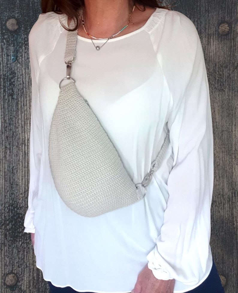 Puede incluir: Un bolso bandolera de crochet gris claro con una correa de cadena plateada. El bolso se lleva sobre el hombro de una persona que lleva una camisa blanca.
