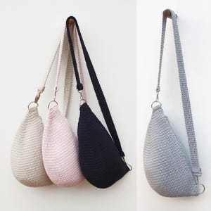 Puede incluir: Tres bolsos de crochet en beige, rosa y negro colgados de una pared blanca. Los bolsos tienen una correa larga y tienen forma de l&aacute;grima.