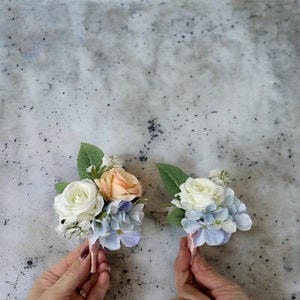 May include: Two small floral arrangements featuring white roses, peach roses, and blue hydrangeas. The arrangements are tied with pink ribbon.