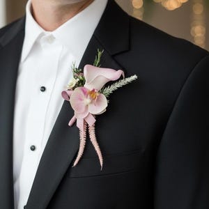 May include: A black tuxedo jacket with a white dress shirt and a pink floral buttonhole. The buttonhole features a pink orchid, calla lily, and fern accents. The jacket has black buttons.
