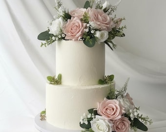 May include: A two-tiered white cake decorated with pink and white roses, greenery, and small white flowers. The cake sits on a white cake stand against a white backdrop.