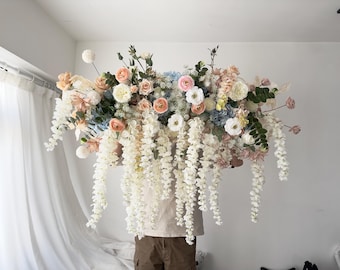May include: A large floral arrangement with white, pink, and blue flowers, greenery, and white cascading flowers. The arrangement is being held by a person.