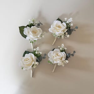 Könnte beinhalten: Vier weiße Rosen-Boutonnières mit Grün und kleinen weißen Blüten.