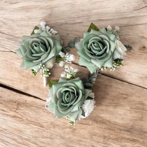 May include: Three sage green rose boutonnieres with white accents and greenery are arranged on a wooden surface. Each boutonniere features a large, fabric rose with detailed petals and small white flower clusters.