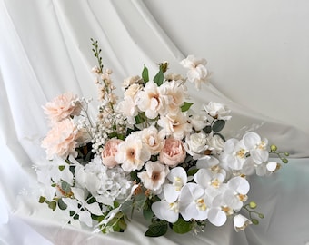 May include: A floral arrangement featuring a variety of flowers in shades of white, cream, and blush pink. The arrangement includes roses, orchids, and other blooms, with green foliage. The flowers are arranged on a white surface.