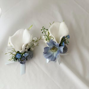 以下が含まれることがあります： 白いカラーの花、青とグレーのアクセントフラワー、緑が特徴の2つのフローラルブートニア。各ブートニアには白いリボンが付いています。背景は白い布です。