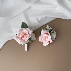 May include: Two artificial boutonnieres with light pink roses, white accent flowers, and green leaves. One has a white ribbon-wrapped stem. The background is a neutral tan surface with white fabric draped across the top.