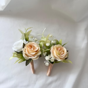 May include: Two boutonnieres with peach-colored roses, white accent flowers, and green leaves. The stems are wrapped in a light peach ribbon. The boutonnieres are set against a white background.