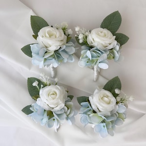 May include: Four floral boutonnieres. Each boutonniere features a white rose, light blue hydrangea, green leaves, and small white accent flowers. The boutonnieres are arranged on a white surface.