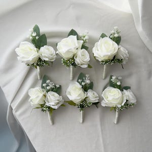 May include: Six white rose boutonnieres with green foliage and small white flowers. Each has a white stem wrapped in white ribbon. The boutonnieres are arranged on a white fabric surface.