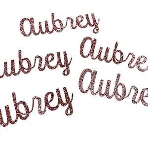 May include: Five rose gold glitter cutouts of the name "Aubrey" in a cursive font.
