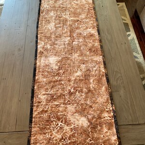 May include: A brown table runner with a white and brown abstract pattern. The runner has a black and gold trim.