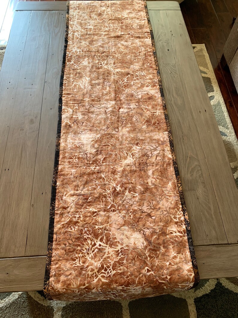 May include: A brown table runner with a white and brown abstract pattern. The runner has a black and gold trim.