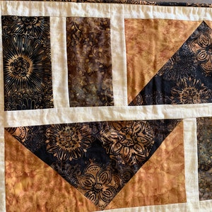 May include: A patchwork quilt table runner with a geometric design. The runner features a black and brown floral pattern, a brown and gold floral pattern, and a solid cream colour.