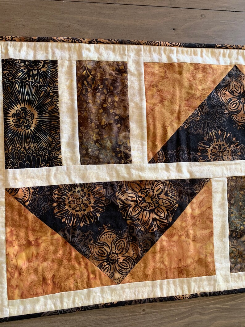 May include: A patchwork quilt table runner with a geometric design. The runner features a black and brown floral pattern, a brown and gold floral pattern, and a solid cream colour.