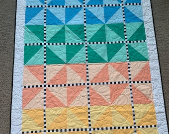 May include: A quilt with a geometric pattern in shades of blue, green, peach, and yellow. The quilt is made up of squares with different patterns within each square. The quilt is edged with a white fabric with black polka dots.
