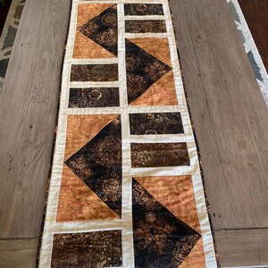 May include: A brown and cream table runner with a geometric pattern. The runner is made of fabric with a batik design.