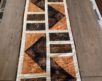 May include: A brown and cream table runner with a geometric pattern. The runner is made of fabric with a batik design.