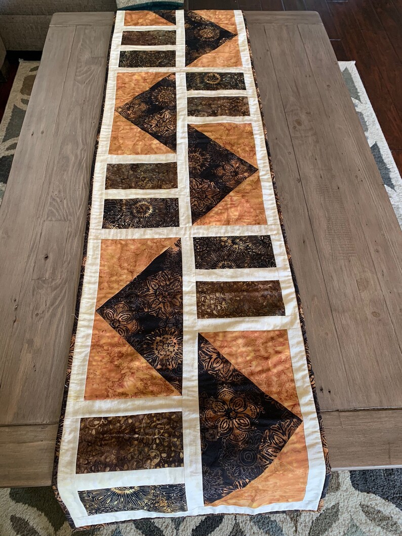 May include: A brown and cream table runner with a geometric pattern. The runner is made of fabric with a batik design.