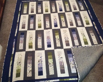 May include: A quilt with a patchwork design featuring a variety of geometric shapes in shades of grey, green, white, and blue. The quilt is bordered with a dark blue fabric and has a grey backing.