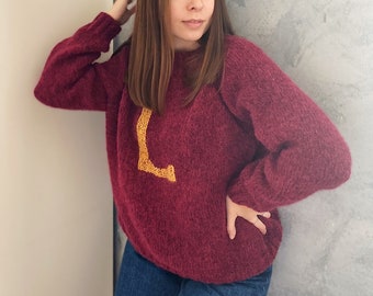 Puede incluir: Una mujer lleva un suéter granate con una letra L dorada en la parte delantera. Lleva pantalones vaqueros azules y tiene el pelo largo y castaño.