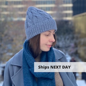 Op de afbeelding: Een blauwe kabelgebreide beanie met een omgeslagen rand. De beanie wordt gedragen door een persoon met lang bruin haar en een blauwe sjaal. De tekst "Ships Next Day" is zichtbaar in de afbeelding.