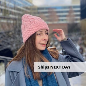 Pode incluir: Um gorro de malha rosa com um design de cabo e uma borda dobrada. O gorro é usado por uma pessoa com cabelo castanho comprido e um casaco azul. O texto "Ships NEXT DAY" é visível na imagem.