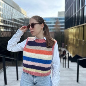 Puede incluir: Una mujer lleva un chaleco de punto a rayas sobre una camisa blanca abotonada y unos vaqueros azules. Lleva gafas de sol y tiene el pelo recogido en una coleta.