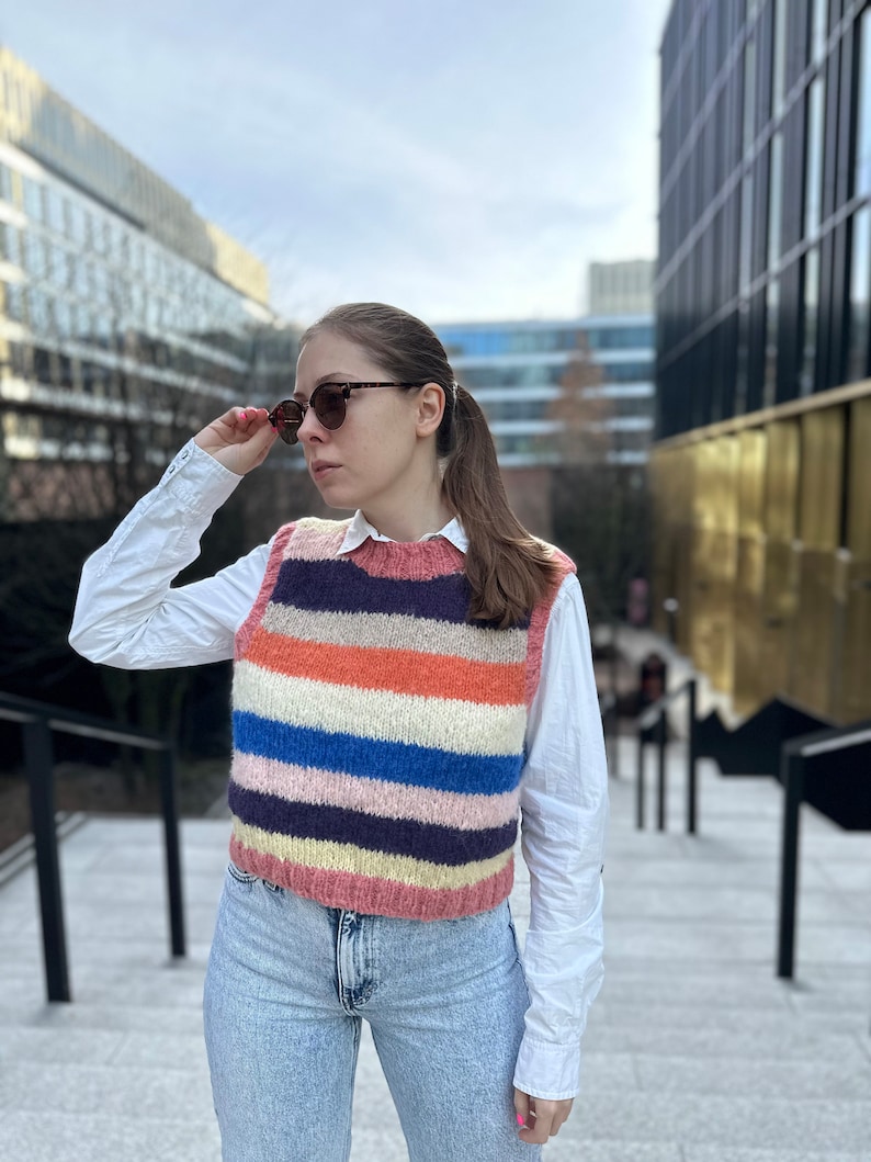 Puede incluir: Una mujer lleva un chaleco de punto a rayas sobre una camisa blanca abotonada y unos vaqueros azules. Lleva gafas de sol y tiene el pelo recogido en una coleta.