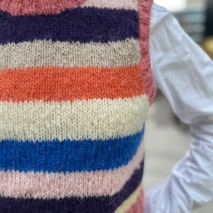 Puede incluir: Un chaleco de punto a rayas con un esquema de color rosa, crema, naranja, azul y morado. El chaleco es sin mangas y tiene cuello redondo.