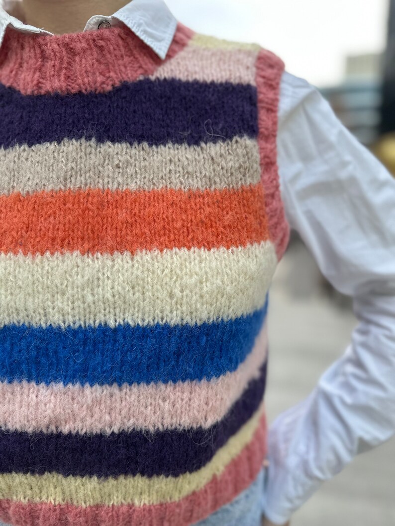 Puede incluir: Un chaleco de punto a rayas con un esquema de color rosa, crema, naranja, azul y morado. El chaleco es sin mangas y tiene cuello redondo.