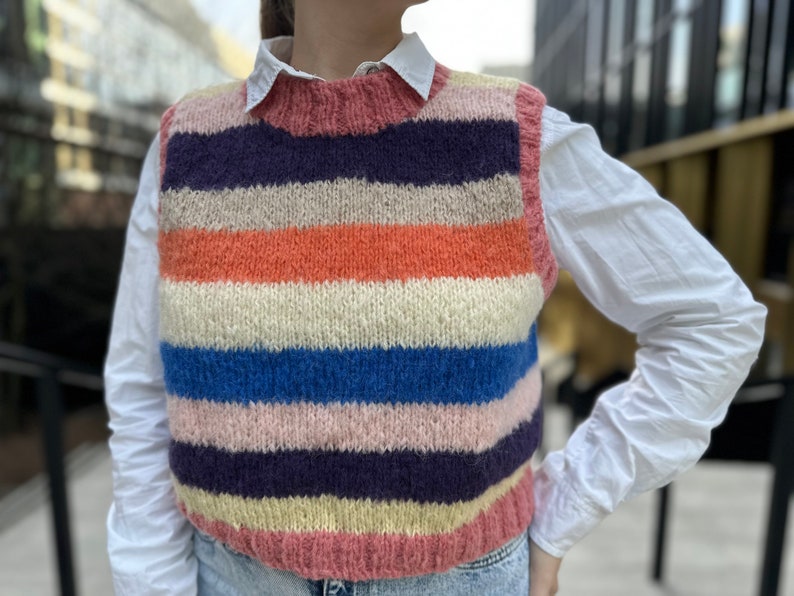 Puede incluir: Un chaleco de punto a rayas con un esquema de color rosa, naranja, azul, blanco y morado. El chaleco es sin mangas y tiene un escote redondo.