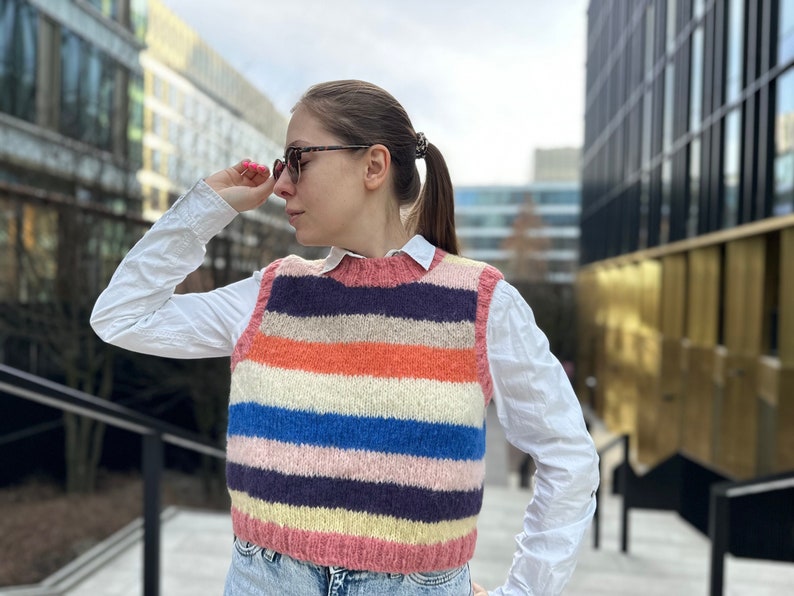 Puede incluir: Una mujer con una camisa blanca y un chaleco de punto de rayas de colores. El chaleco tiene rayas horizontales en tonos de rosa, naranja, azul y blanco.