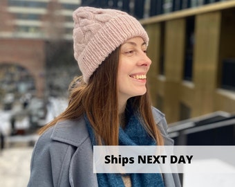 Könnte beinhalten: Eine Frau trägt eine rosafarbene Strickmütze mit Zopfmuster und einen blauen Schal. Der Text "Ships Next Day" ist im Bild sichtbar.