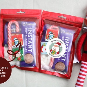 May include: Two red bags filled with a Cadbury hot chocolate packet, candy cane, marshmallows, and a small Santa sticker. The bags are next to a red and white striped elf doll.