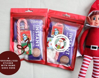 May include: Two red bags filled with a Cadbury hot chocolate packet, candy cane, marshmallows, and a small Santa sticker. The bags are next to a red and white striped elf doll.