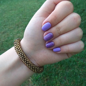 May include: A close-up of a hand with purple nail polish, including one glitter-covered nail. A gold beaded bracelet is on the wrist. The background is blurred green grass.