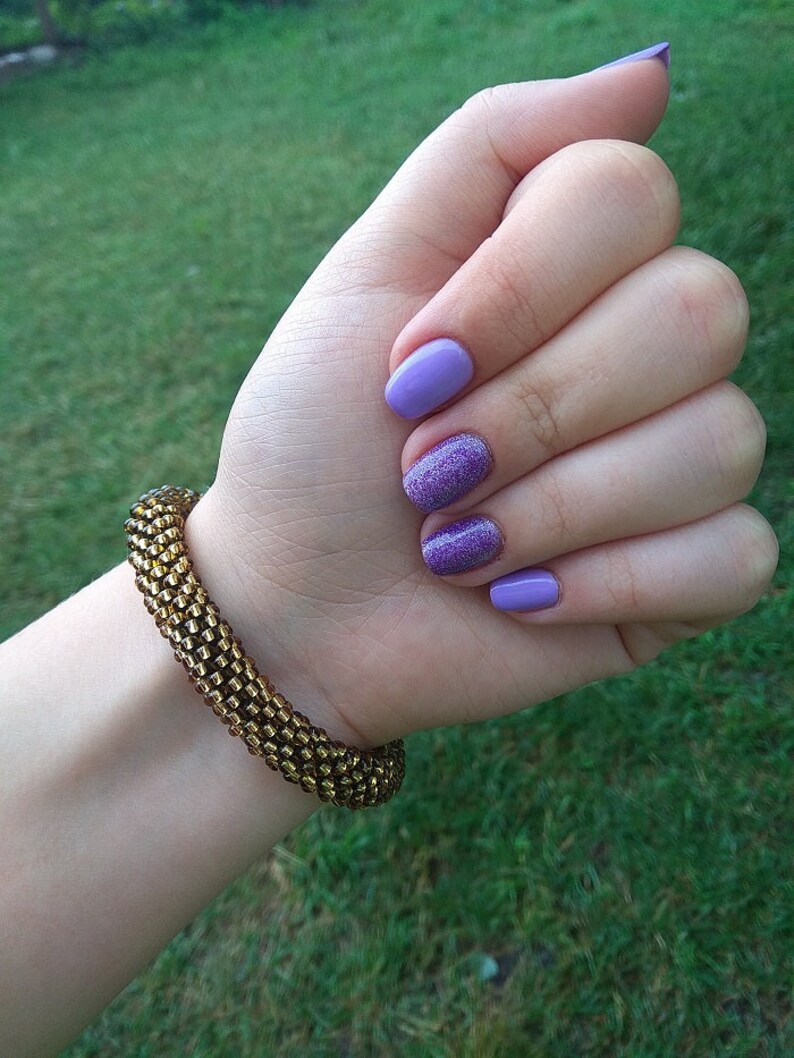 May include: A close-up of a hand with purple nail polish, including one glitter-covered nail. A gold beaded bracelet is on the wrist. The background is blurred green grass.